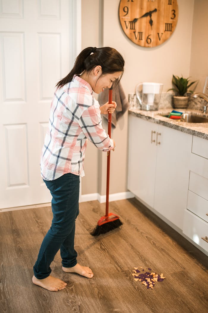 Asian woman cleaning kitchen with a broom, demonstrating domestic chores in a casual setting.