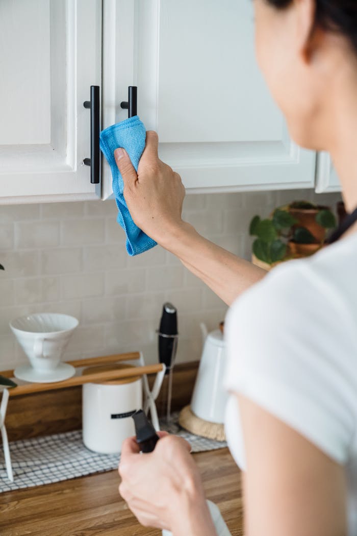 A person cleaning a white kitchen cabinet using a blue cloth and spray bottle.